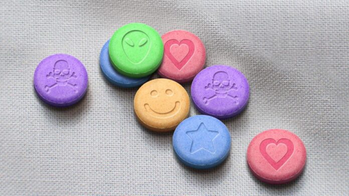 Na obrazie znajduje się kilka kolorowych tabletek MDMA o różnych kształtach i rozmiarach, rozłożonych na szarym tkaninowym tle. Tabletki mają różne kolory: zielony, różowy, fioletowy, niebieski i beżowy. Każda z nich jest ozdobiona innym symbolem: uśmiechniętą twarzą, sercem, gwiazdą oraz czaszką z kośćmi.