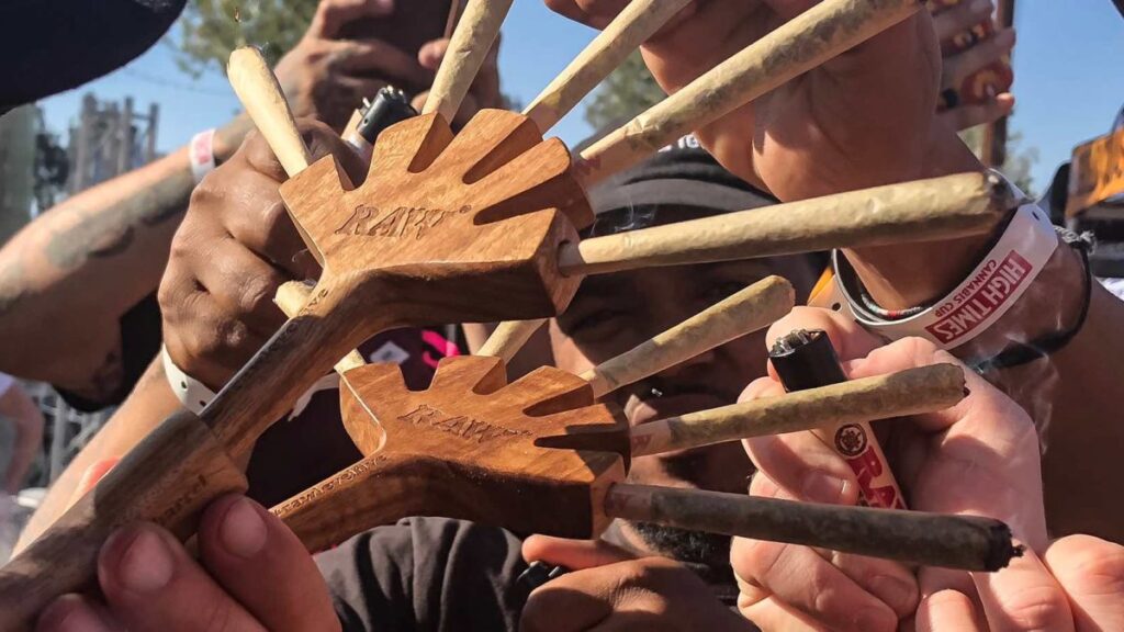 Drewniany holder na jointy marki RAW z wypalonym logo, trzymany przez osoby w tłumie. Holder ma kształt ręki z rozstawionymi palcami, w których umieszczono kilka jointów, gotowych do zapalenia