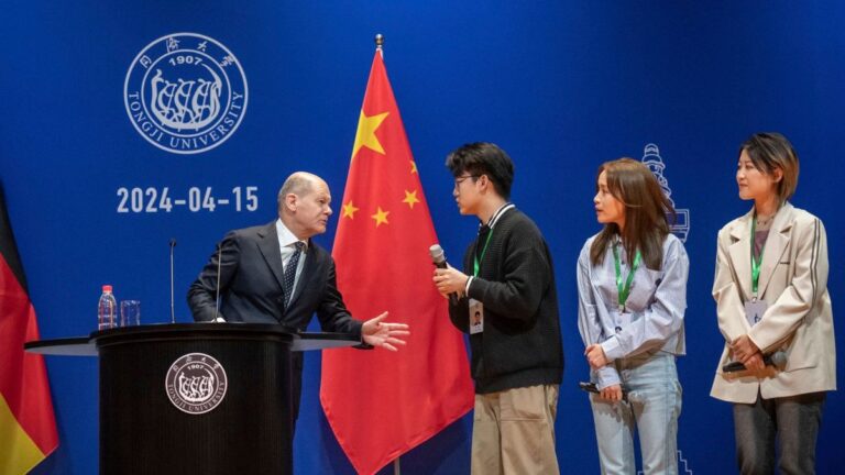Kanclerz Niemiec Olaf Scholz stoi przy mównicy w sali wykładowej Uniwersytetu Tongji, obok niego stoi chińska flaga. Naprzeciwko niego znajduje się młody mężczyzna trzymający mikrofon, wydaje się zadawać pytanie. Obok studenta stoją dwie młode kobiety, również ze znacznikami identyfikacyjnymi, które wydają się czekać na swoją kolej. Atmosfera jest formalna, a uczestnicy są skupieni na wymianie dialogu.