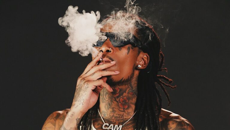 Wiz Khalifa, amerykański raper, w ciemnych okularach i z licznymi tatuażami na ciele, pali marihuanę, tworząc chmurę dymu.